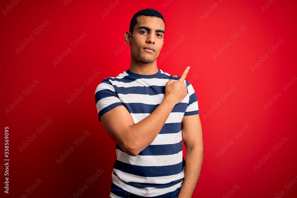Fototapeta premium Handsome african american man wearing casual striped t-shirt standing over red background Pointing with hand finger to the side showing advertisement, serious and calm face
