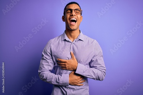 Handsome african american man wearing striped shirt and glasses over purple background smiling and laughing hard out loud because funny crazy joke with hands on body.
