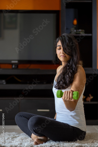 woman doing weights with one arm sitting at home in her room for quarantine and coronavirus pandemic in mexico, wearing white sportswear and black pants