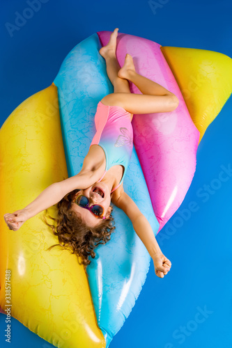 Pretty smiling girl wearing pink and blue swimwear and sunglasses lies, relax on rainbow inflatable mattress. Top view