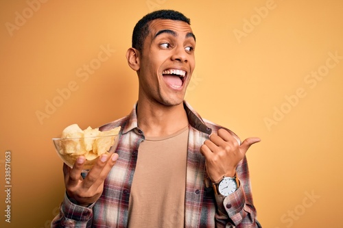 Young handsome african american man holding bowl of chips potatoes over yellow background pointing and showing with thumb up to the side with happy face smiling