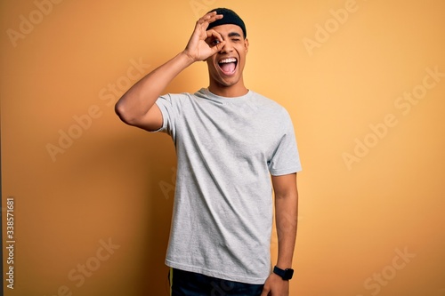 Handsome african american sportsman doing sport wearing sportswear over yellow background doing ok gesture with hand smiling, eye looking through fingers with happy face. © Krakenimages.com