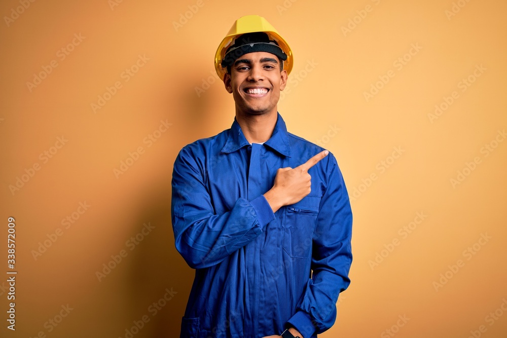 Young handsome african american worker man wearing blue uniform and security helmet cheerful with a smile on face pointing with hand and finger up to the side with happy and natural expression