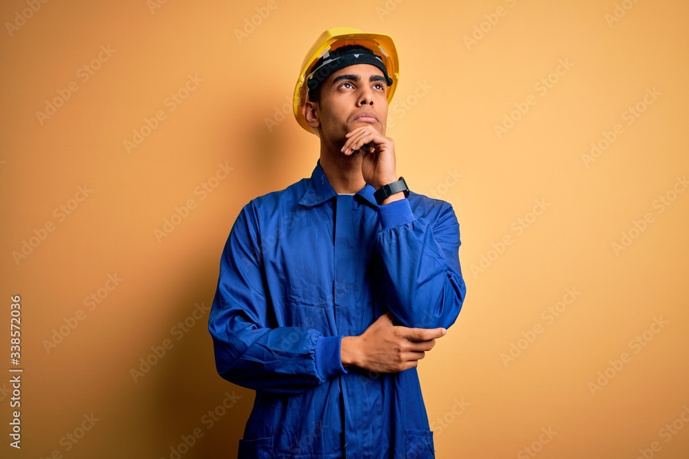 Young handsome african american worker man wearing blue uniform and security helmet with hand on chin thinking about question, pensive expression. Smiling and thoughtful face. Doubt concept.