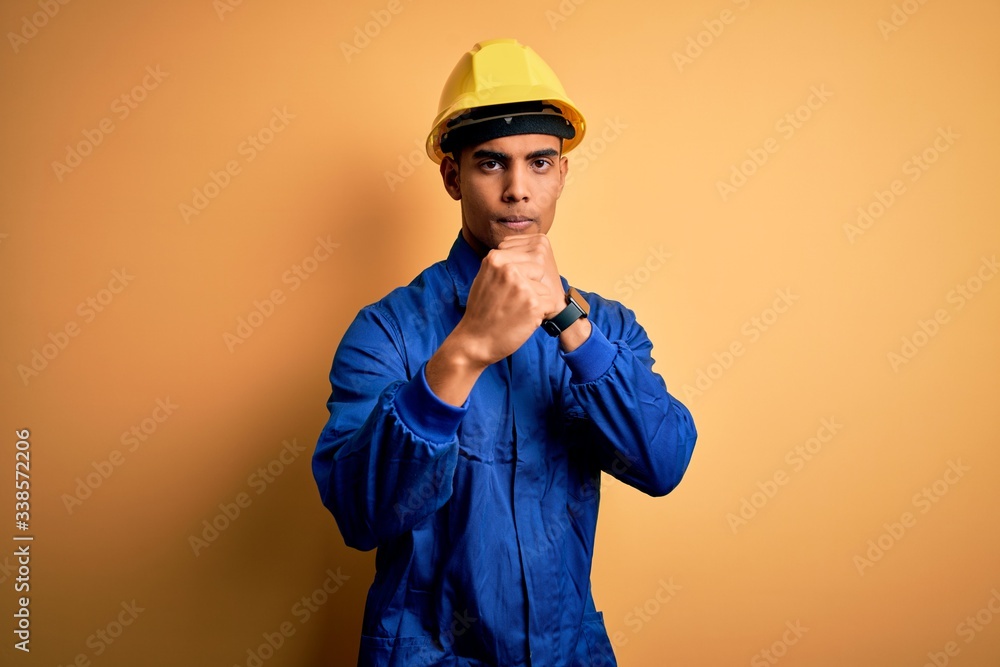 Young handsome african american worker man wearing blue uniform and security helmet Ready to fight with fist defense gesture, angry and upset face, afraid of problem