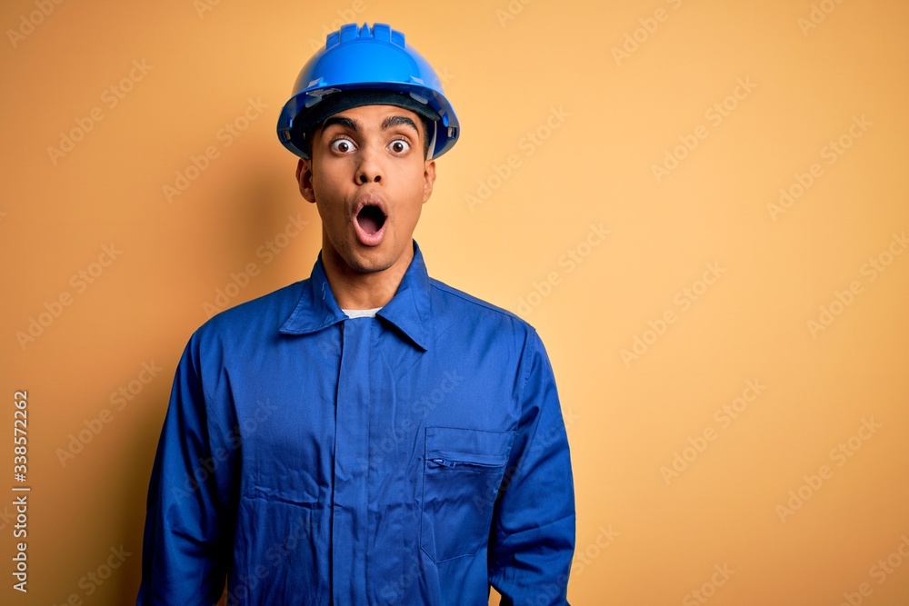 Young handsome african american worker man wearing blue uniform and security helmet afraid and shocked with surprise expression, fear and excited face. Young handsome african american worker man wearing blue uniform and security helmet afraid and shocked with surprise expression, fear and excited face.