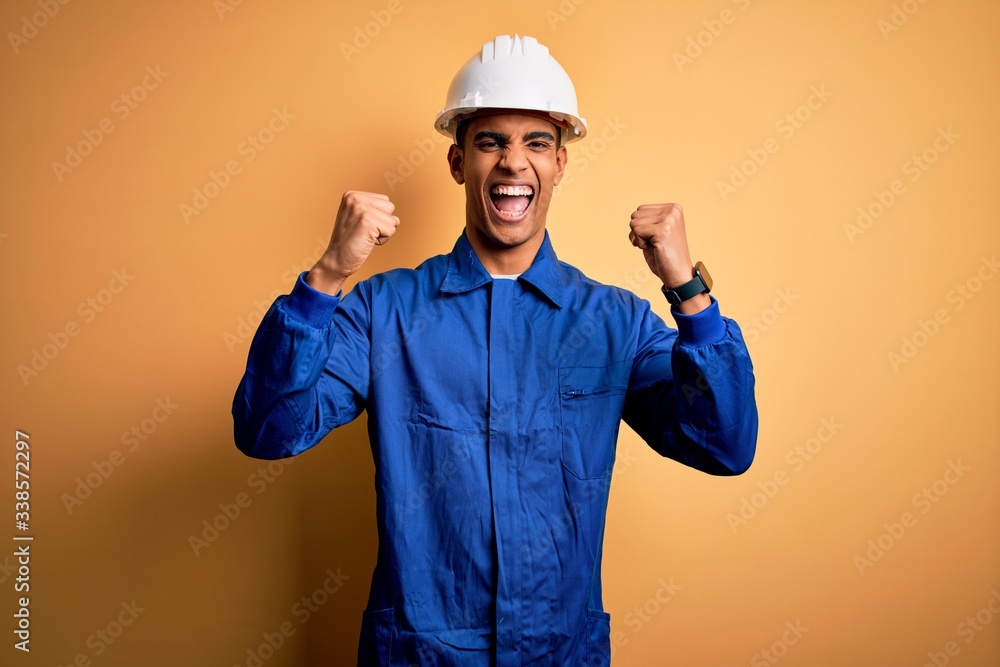 Young handsome african american worker man wearing blue uniform and security helmet celebrating surprised and amazed for success with arms raised and open eyes. Winner concept.