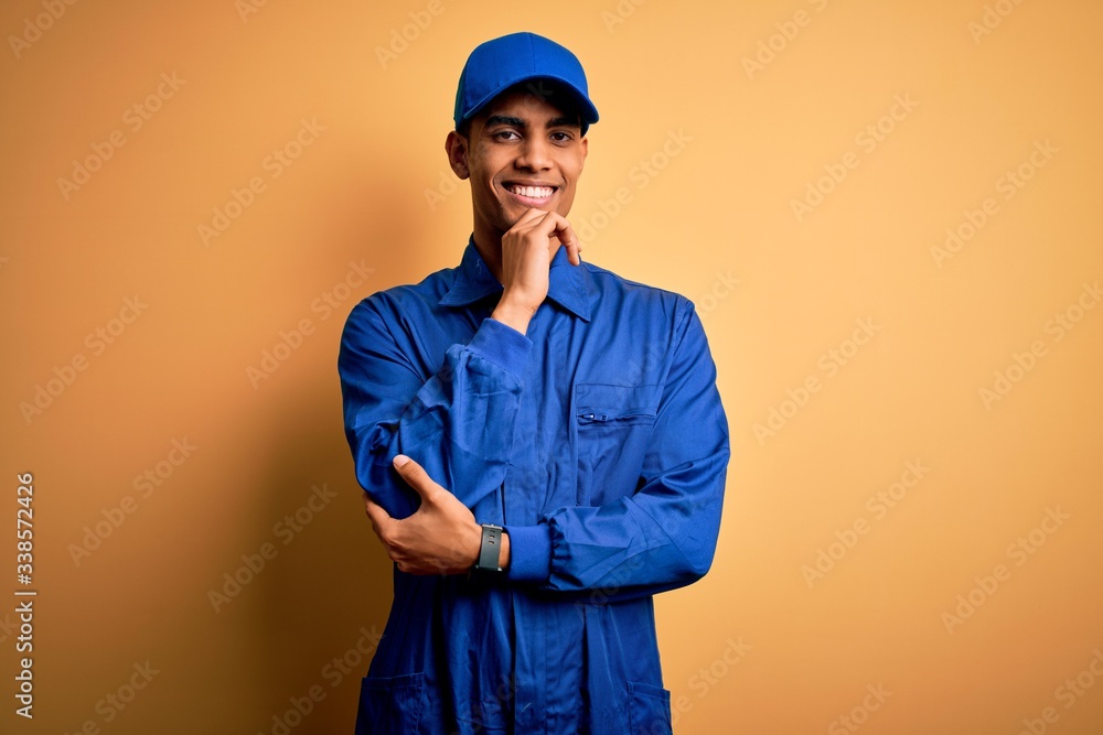 Young african american mechanic man wearing blue uniform and cap over yellow background looking confident at the camera with smile with crossed arms and hand raised on chin. Thinking positive.