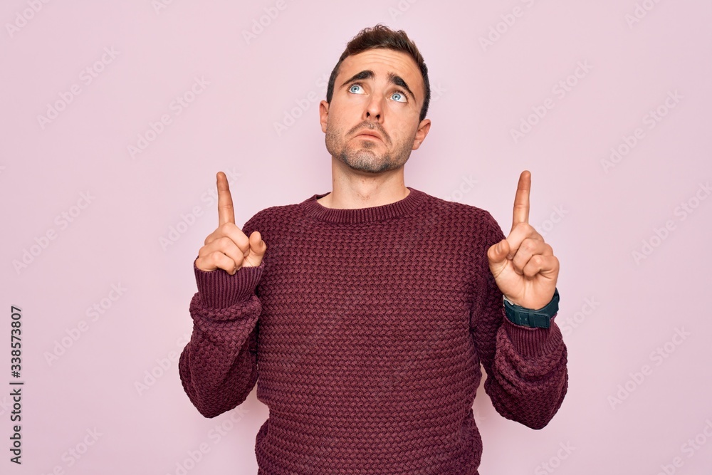Young handsome man with blue eyes wearing casual sweater standing over pink background Pointing up looking sad and upset, indicating direction with fingers, unhappy and depressed. Young handsome man with blue eyes wearing casual sweater standing over pink background Pointing up looking sad and upset, indicating direction with fingers, unhappy and depressed.