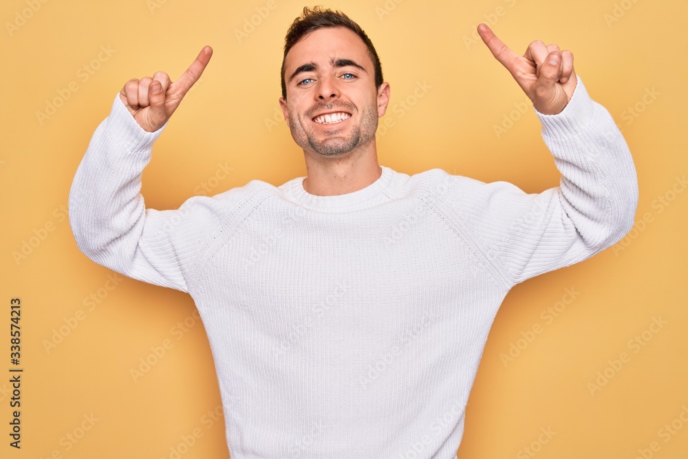 Young handsome man with blue eyes wearing casual sweater standing over yellow background smiling amazed and surprised and pointing up with fingers and raised arms. Young handsome man with blue eyes wearing casual sweater standing over yellow background smiling amazed and surprised and pointing up with fingers and raised arms.