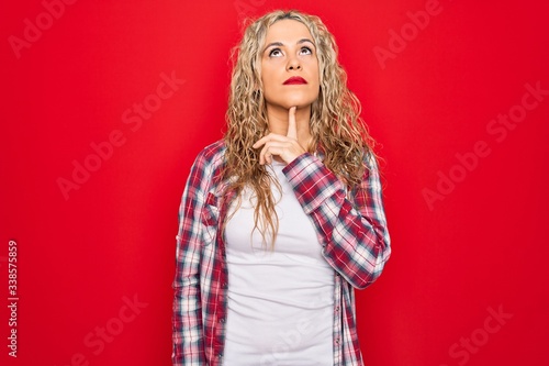 Young beautiful blonde woman wearing casual shirt standing over isolated red background Thinking concentrated about doubt with finger on chin and looking up wondering