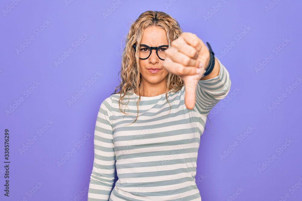 Beautiful blonde woman wearing casual striped t-shirt and glasses over purple background looking unhappy and angry showing rejection and negative with thumbs down gesture. Bad expression.
