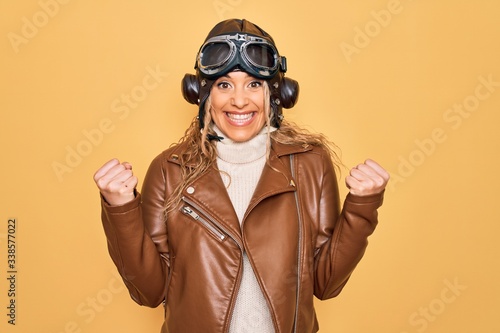 Fotografia Young beautiful blonde aviator woman wearing vintage pilot helmet whit glasses and jacket celebrating surprised and amazed for success with arms raised and open eyes