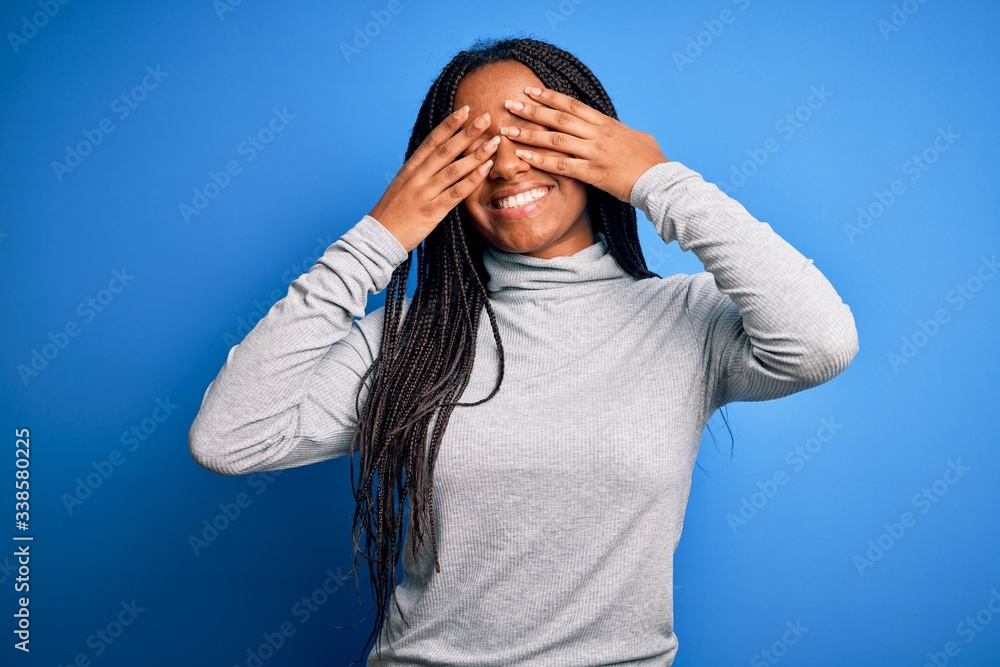 Young african american woman standing wearing casual turtleneck over blue isolated background covering eyes with hands smiling cheerful and funny. Blind concept.