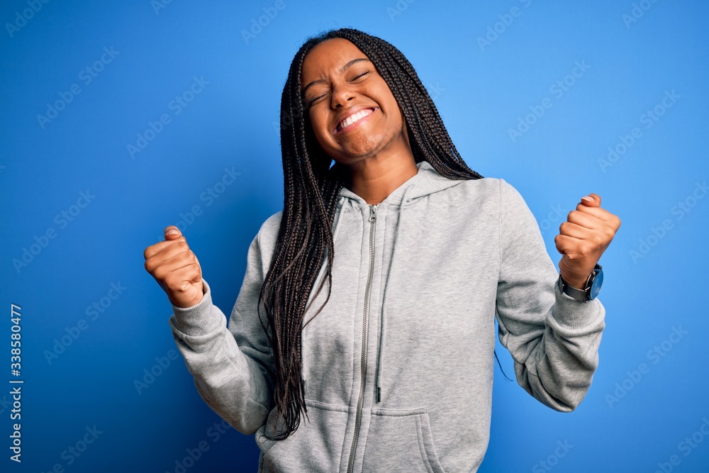 Young african american athlete woman wearing sports sweatshirt over blue isolated background very happy and excited doing winner gesture with arms raised, smiling and screaming for success. Celebrate Young african american athlete woman wearing sports sweatshirt over blue isolated background very happy and excited doing winner gesture with arms raised, smiling and screaming for success. Celebrate