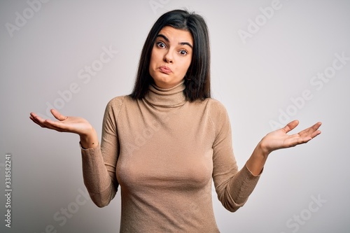 Fotografering Young beautiful brunette woman wearing turtleneck sweater over white background clueless and confused expression with arms and hands raised