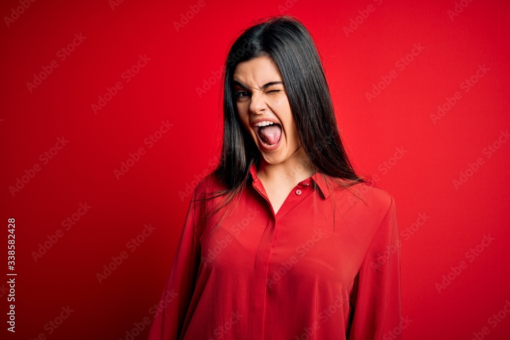 Young beautiful brunette woman wearing casual shirt standing over red background winking looking at the camera with sexy expression, cheerful and happy face. Young beautiful brunette woman wearing casual shirt standing over red background winking looking at the camera with sexy expression, cheerful and happy face.