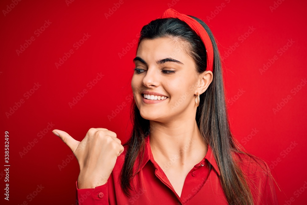 Young beautiful brunette woman wearing casual shirt and diadem over red background smiling with happy face looking and pointing to the side with thumb up. Young beautiful brunette woman wearing casual shirt and diadem over red background smiling with happy face looking and pointing to the side with thumb up.
