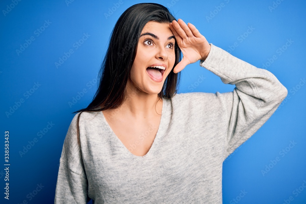 Young beautiful brunette woman wearing casual sweater standing over blue background very happy and smiling looking far away with hand over head. Searching concept. Young beautiful brunette woman wearing casual sweater standing over blue background very happy and smiling looking far away with hand over head. Searching concept.