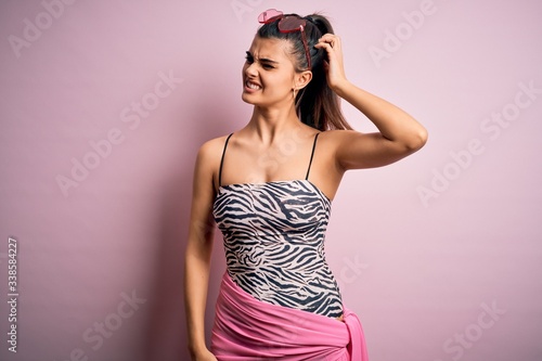 Young beautiful brunette woman on vacation wearing swimsuit over pink background confuse and wondering about question. Uncertain with doubt, thinking with hand on head. Pensive concept.