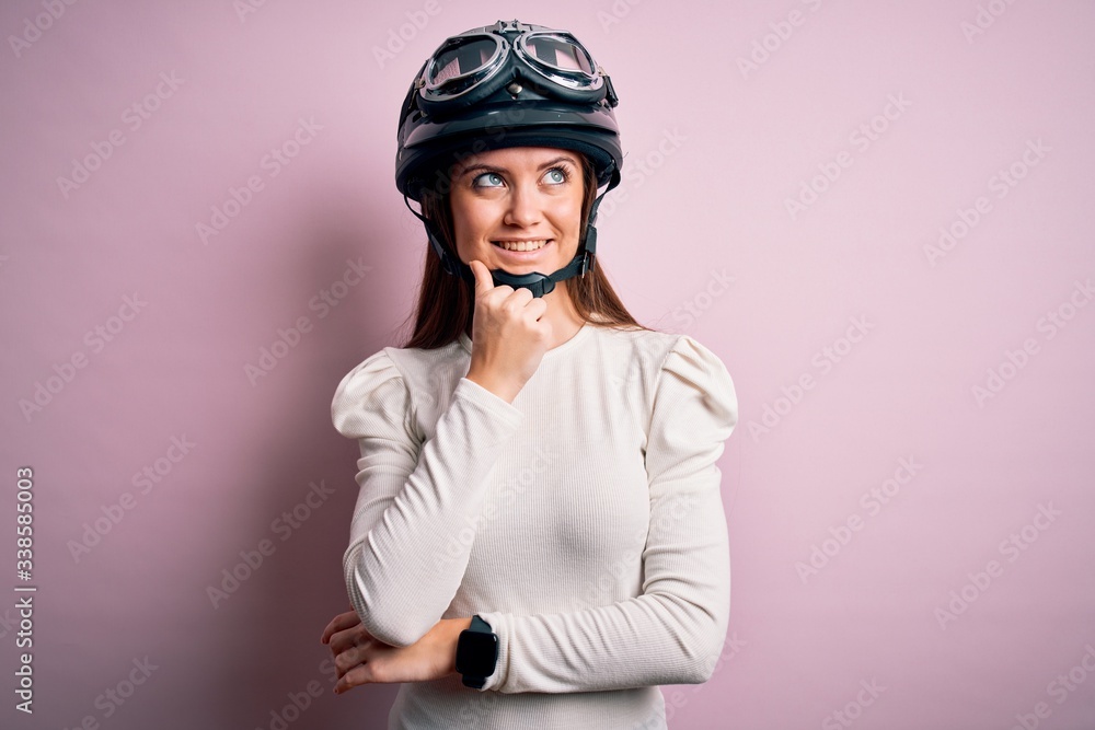 Young beautiful motorcyclist woman with blue eyes wearing moto helmet over pink background with hand on chin thinking about question, pensive expression. Smiling and thoughtful face. Doubt concept.