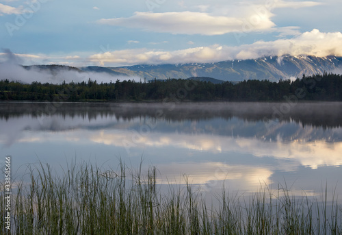 Morning. Dawn. Sky and clouds are reflected on the surface of the water. In the distance, the opposite shore of the lake and a mountain range are visible.