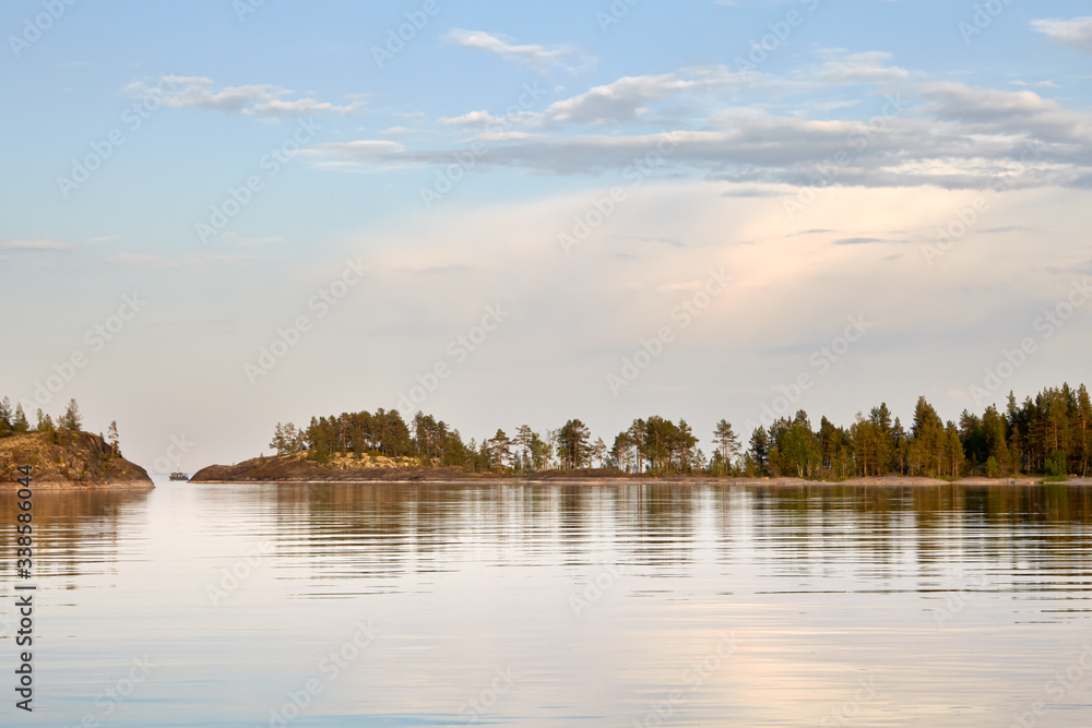 Summer. Evening.Calm water surface of the lake. In the distance you can see a narrow strip of rocky shore,overgrown with moss and trees.On the left is the passage between the Islands. Gentle blue sky.