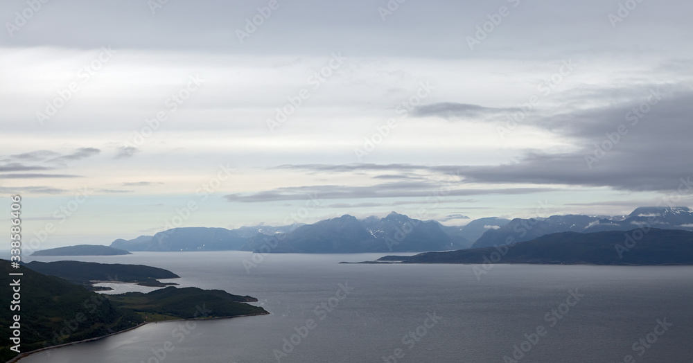 Norway. Summer. Evening. Bay with dark-gray water goes into the distance. On the left is visible part of a small island. In the background are the ridges of the fjord. A light gray sky with clouds.