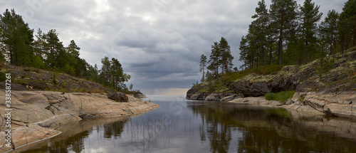 Summer. In the foreground we see the Bay of lake Ladoga  surrounded on the left and right by rocky shores. The upper part of the banks is covered with pine trees.Quiet  calm water. A dark  stormy sky.