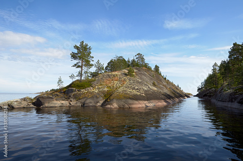 Lake. Two large stone islands protrude from the water. Between them is a narrow long duct. The islands are covered with moss, shrubs and trees. Light blue sky with clouds. Summer. Russia.