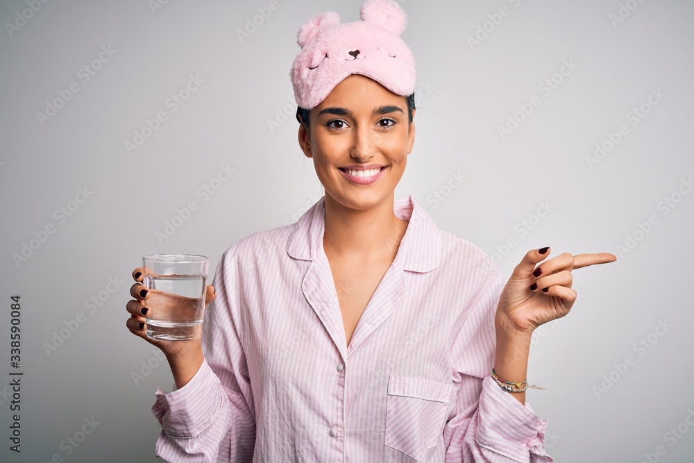 Young beautiful woman wearing pajama and sleep mask drinking glass of water very happy pointing with hand and finger to the side Young beautiful woman wearing pajama and sleep mask drinking glass of water very happy pointing with hand and finger to the side