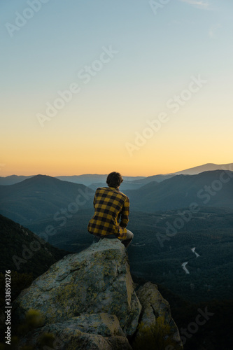Young boy sitting on rocks watching a mountain landscape, with an incredible sunset, warm sky with clouds. Vertical image. © nil.fort