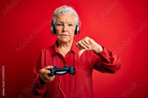 Senior beautiful grey-haired gamer woman playing video game using joystick and headphones with angry face, negative sign showing dislike with thumbs down, rejection concept