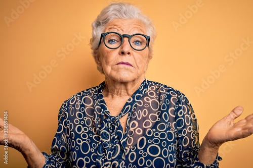 Senior beautiful grey-haired woman wearing casual shirt and glasses over yellow background clueless and confused expression with arms and hands raised. Doubt concept.