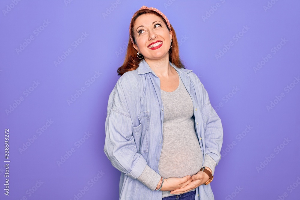 Young beautiful redhead pregnant woman expecting baby over isolated purple background looking away to side with smile on face, natural expression. Laughing confident. Young beautiful redhead pregnant woman expecting baby over isolated purple background looking away to side with smile on face, natural expression. Laughing confident.