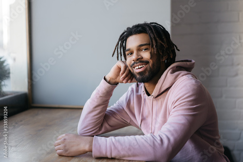Portrait of happy African American man with stylish dreadlocks looking at camera and smiling, sitting in cafe 