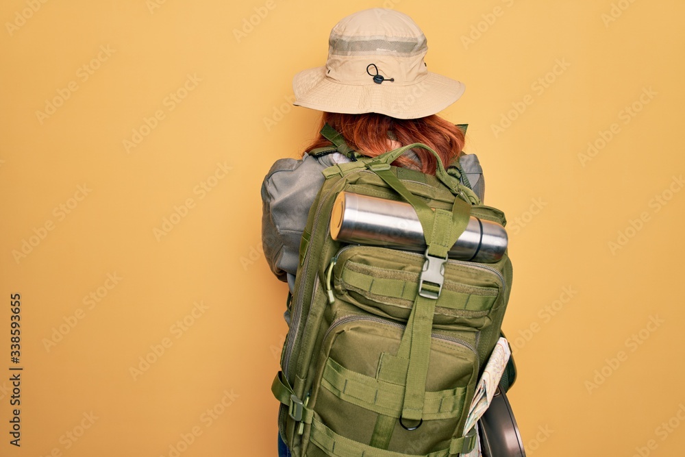 Young redhead backpacker woman hiking wearing backpack and hat over yellow background standing backwards looking away with crossed arms Young redhead backpacker woman hiking wearing backpack and hat over yellow background standing backwards looking away with crossed arms