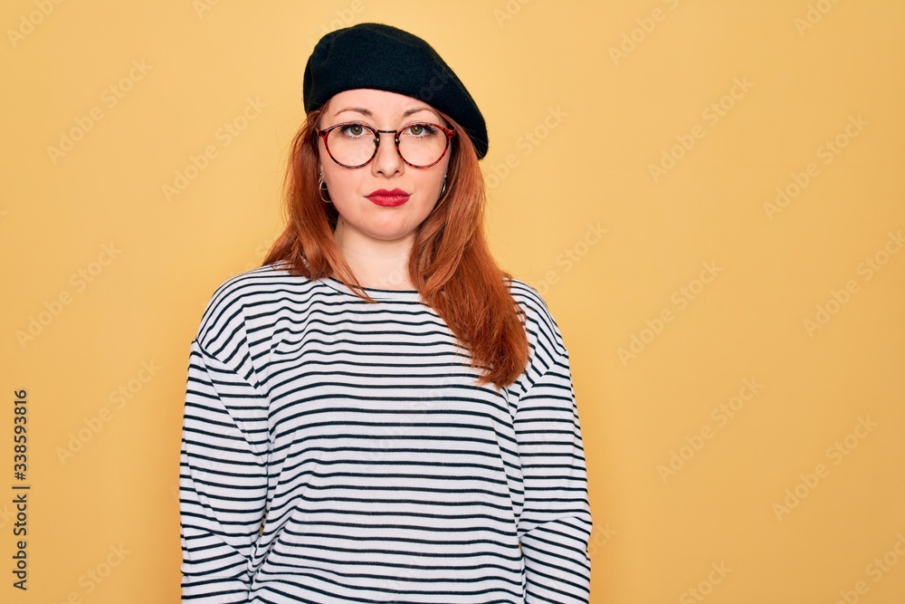 Beautiful redhead woman wearing striped t-shirt and french beret over yellow background with serious expression on face. Simple and natural looking at the camera. Beautiful redhead woman wearing striped t-shirt and french beret over yellow background with serious expression on face. Simple and natural looking at the camera.