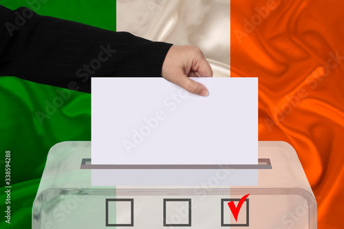 female voter drops a ballot in a transparent ballot box against the background of the national flag of Ireland, concept of state elections, referendum