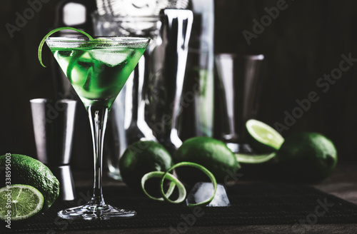 Green alcoholic cocktail martini glass with dry gin, vermouth, liquor, lime zest and ice, bar tools, dark background