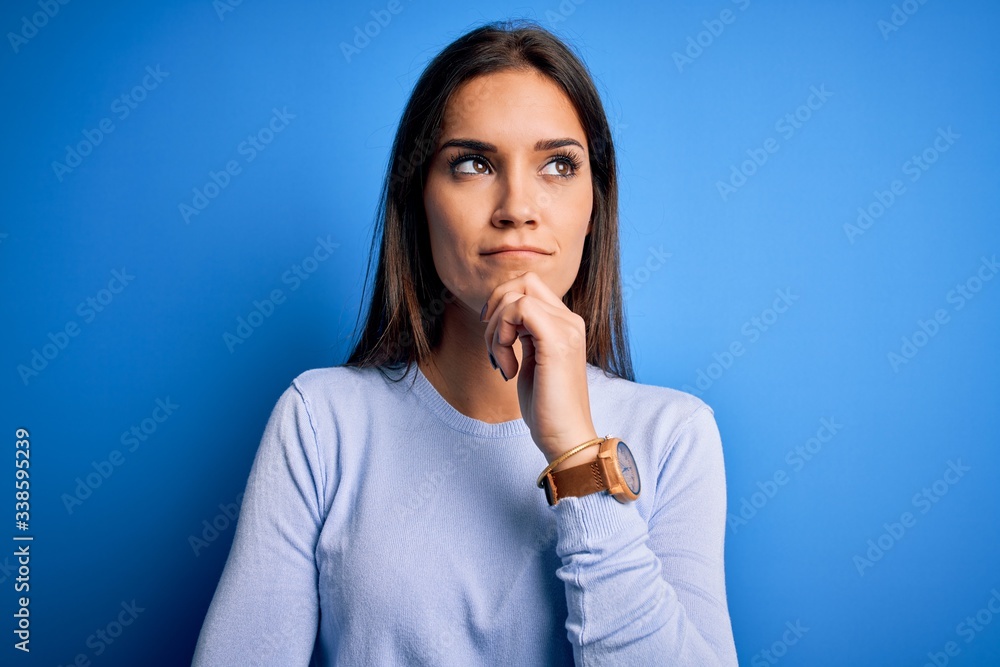 Young beautiful brunette woman wearing casual sweater standing over blue background with hand on chin thinking about question, pensive expression. Smiling and thoughtful face. Doubt concept. Young beautiful brunette woman wearing casual sweater standing over blue background with hand on chin thinking about question, pensive expression. Smiling and thoughtful face. Doubt concept.