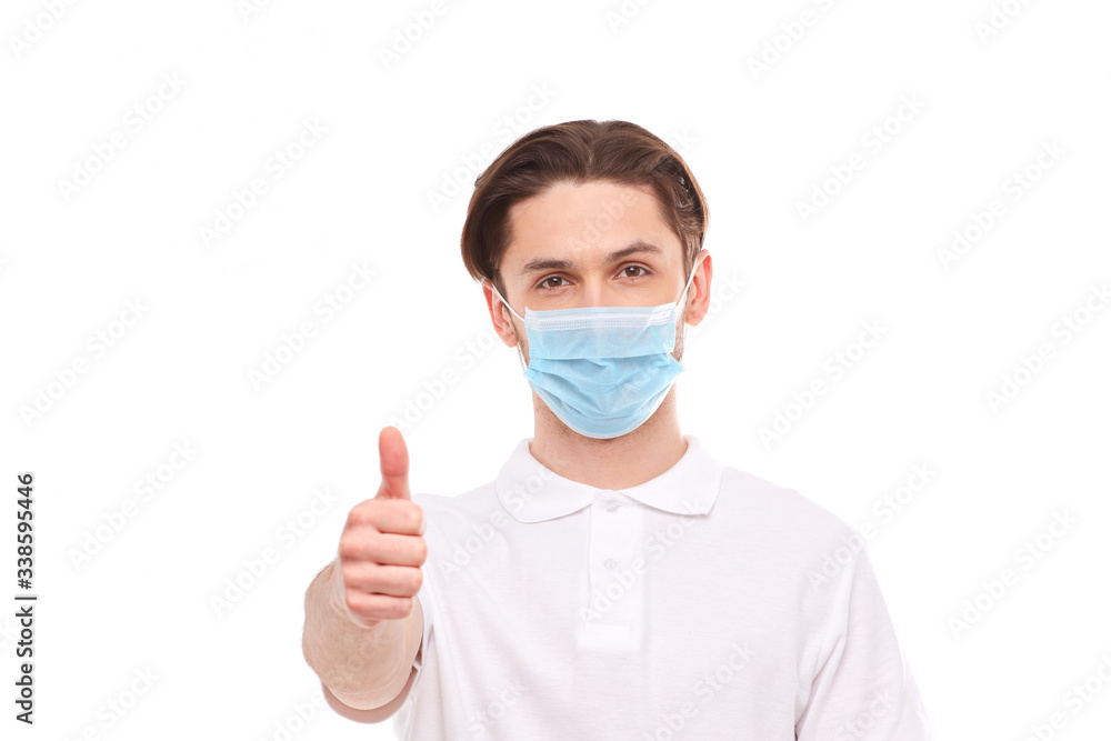 A man in a white T-shirt of European appearance in a medical mask, shows a thumb up. In the mask you are protected from covid 19 viruses.