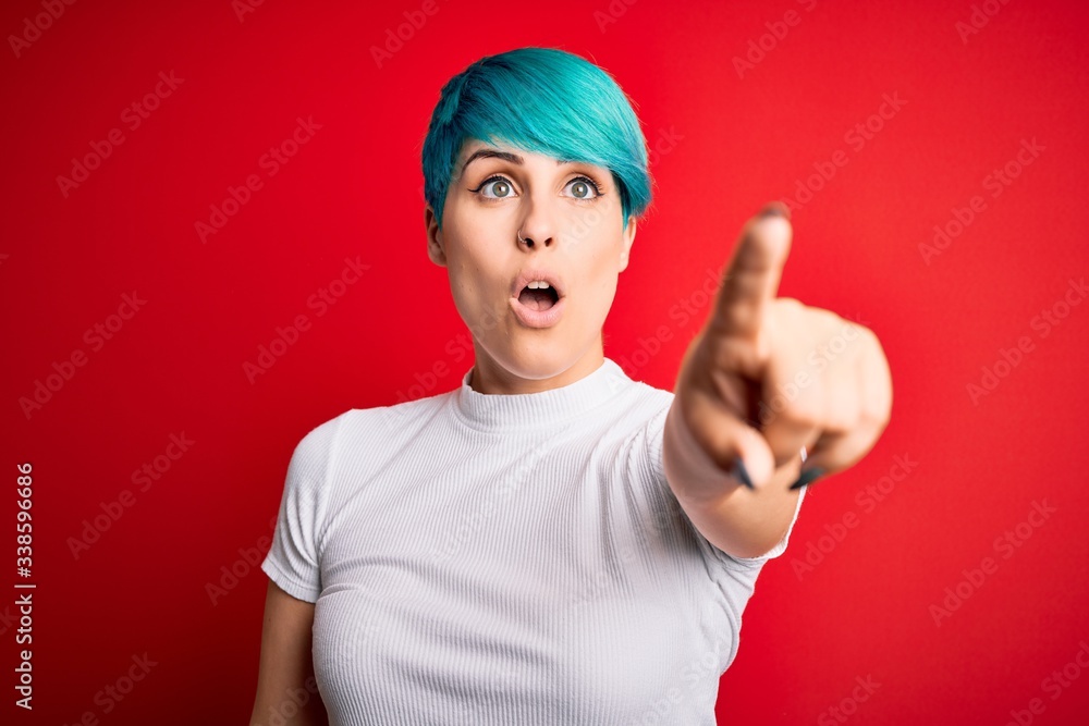Young beautiful woman with blue fashion hair wearing casual t-shirt over red background Pointing with finger surprised ahead, open mouth amazed expression, something on the front Young beautiful woman with blue fashion hair wearing casual t-shirt over red background Pointing with finger surprised ahead, open mouth amazed expression, something on the front