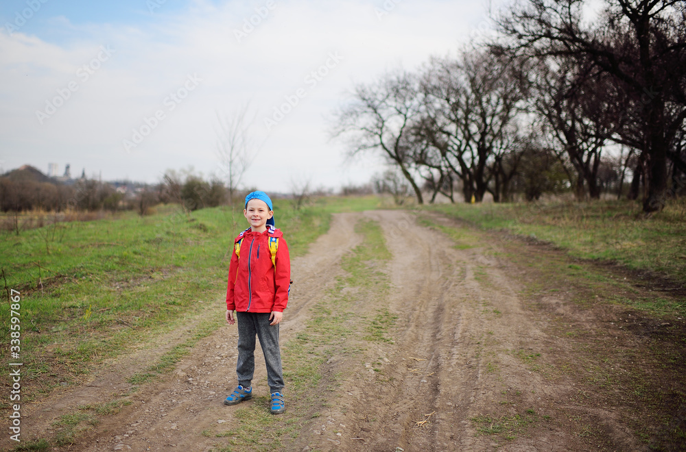 a small happy child a boy in a red jacket and a blue cap stands and smiles against the background of a spring landscape in a Park or forest. Space for text, copy space
