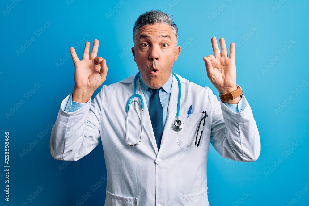 Middle age handsome grey-haired doctor man wearing coat and blue stethoscope looking surprised and shocked doing ok approval symbol with fingers. Crazy expression Middle age handsome grey-haired doctor man wearing coat and blue stethoscope looking surprised and shocked doing ok approval symbol with fingers. Crazy expression