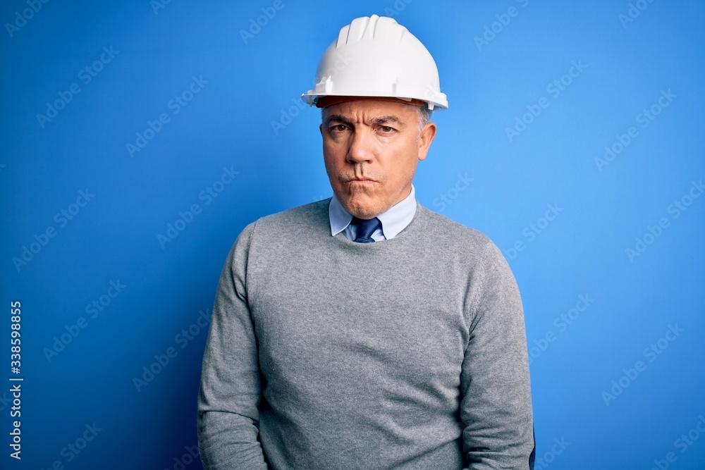 Middle age handsome grey-haired engineer man wearing safety helmet over blue background skeptic and nervous, frowning upset because of problem. Negative person.