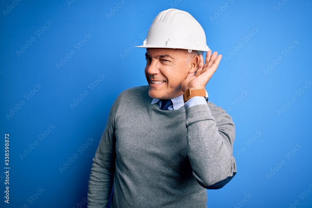 Middle age handsome grey-haired engineer man wearing safety helmet over blue background smiling with hand over ear listening an hearing to rumor or gossip. Deafness concept.