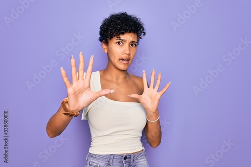 Young beautiful african american afro woman wearing casual t-shirt over purple background afraid and terrified with fear expression stop gesture with hands, shouting in shock. Panic concept.
