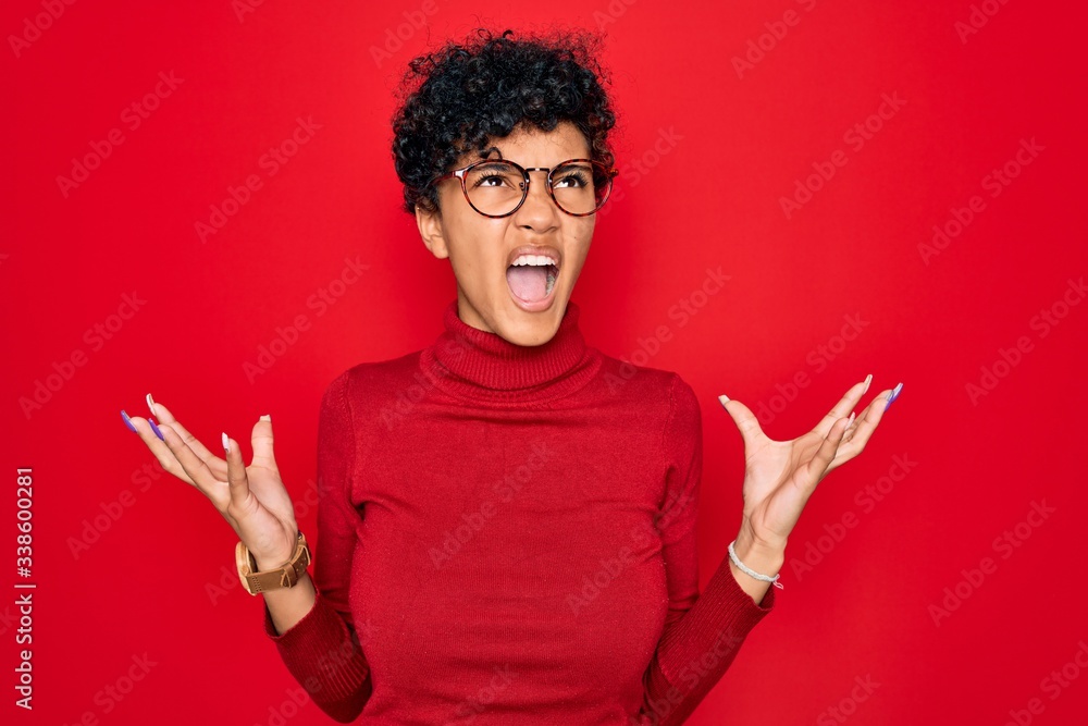 Young beautiful african american afro woman wearing turtleneck sweater and glasses crazy and mad shouting and yelling with aggressive expression and arms raised. Frustration concept. Young beautiful african american afro woman wearing turtleneck sweater and glasses crazy and mad shouting and yelling with aggressive expression and arms raised. Frustration concept.
