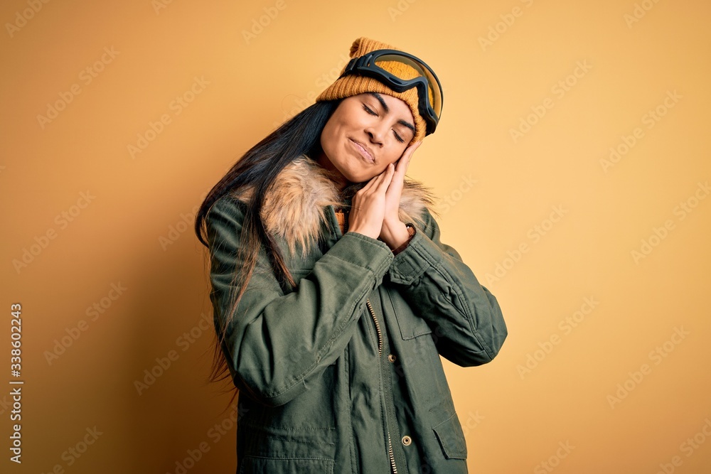 Young beautiful hispanic woman wearing ski glasses and coat for winter weather sleeping tired dreaming and posing with hands together while smiling with closed eyes. Young beautiful hispanic woman wearing ski glasses and coat for winter weather sleeping tired dreaming and posing with hands together while smiling with closed eyes.
